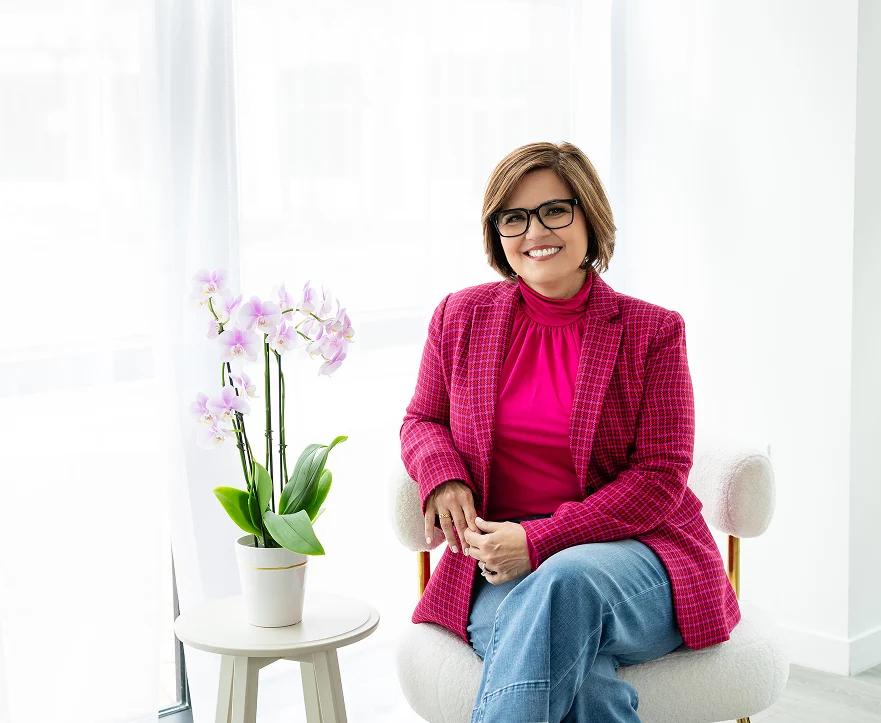 Joni Marmo seated in a bright room, smiling in a bold pink jacket beside a blooming orchid, ready to share wisdom and encouragement.